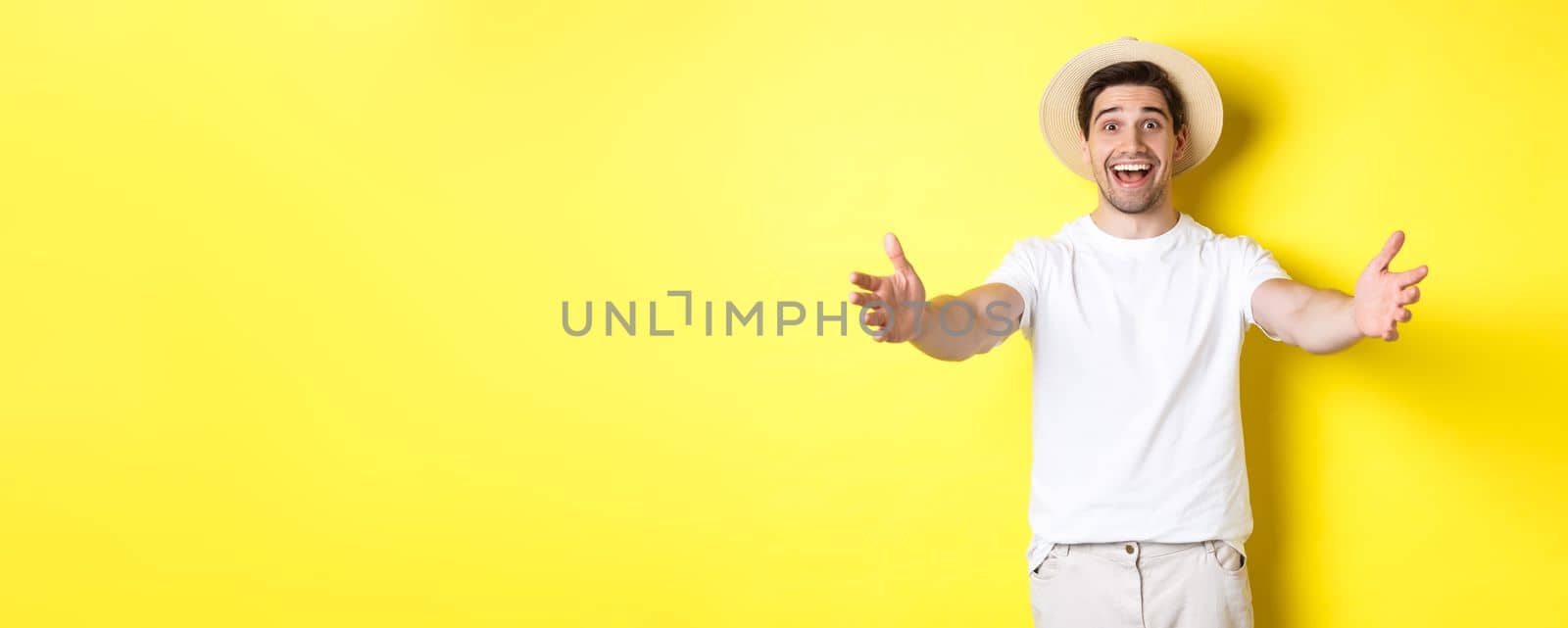 Concept of tourism and summer. Happy and friendly man traveller reaching hands for hug, greeting or welcome you, standing over yellow background.