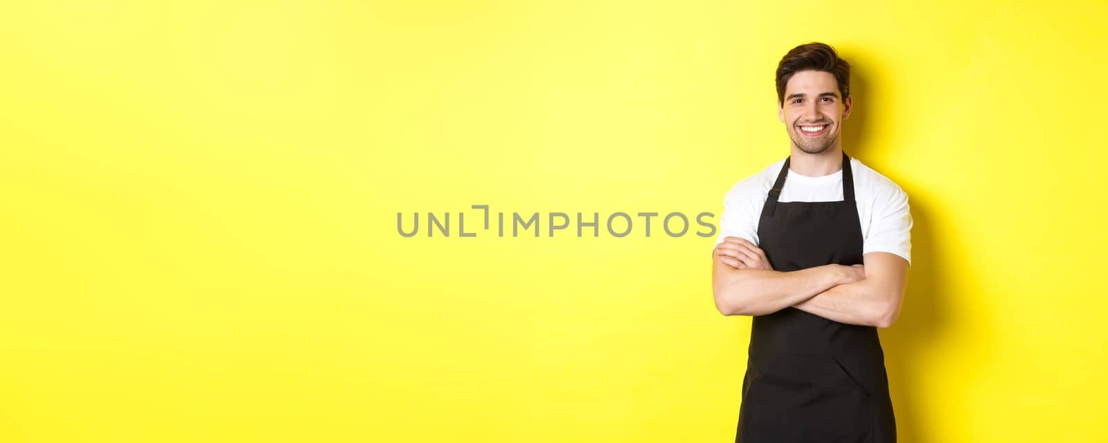 Smiling male waiter in black apron standing confident, cross arms on chest against yellow background.