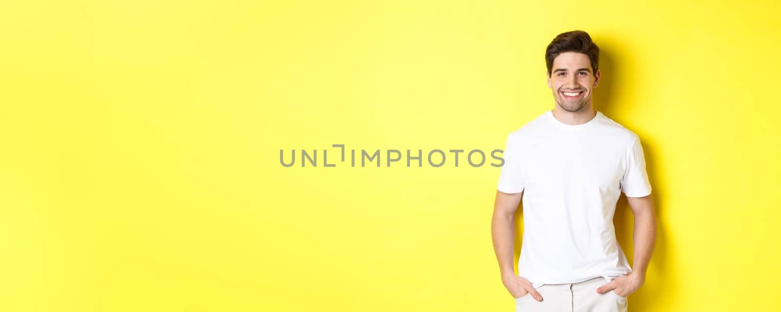 Young handsome man smiling at camera, holding hands in pockets, standing against yellow background by Benzoix