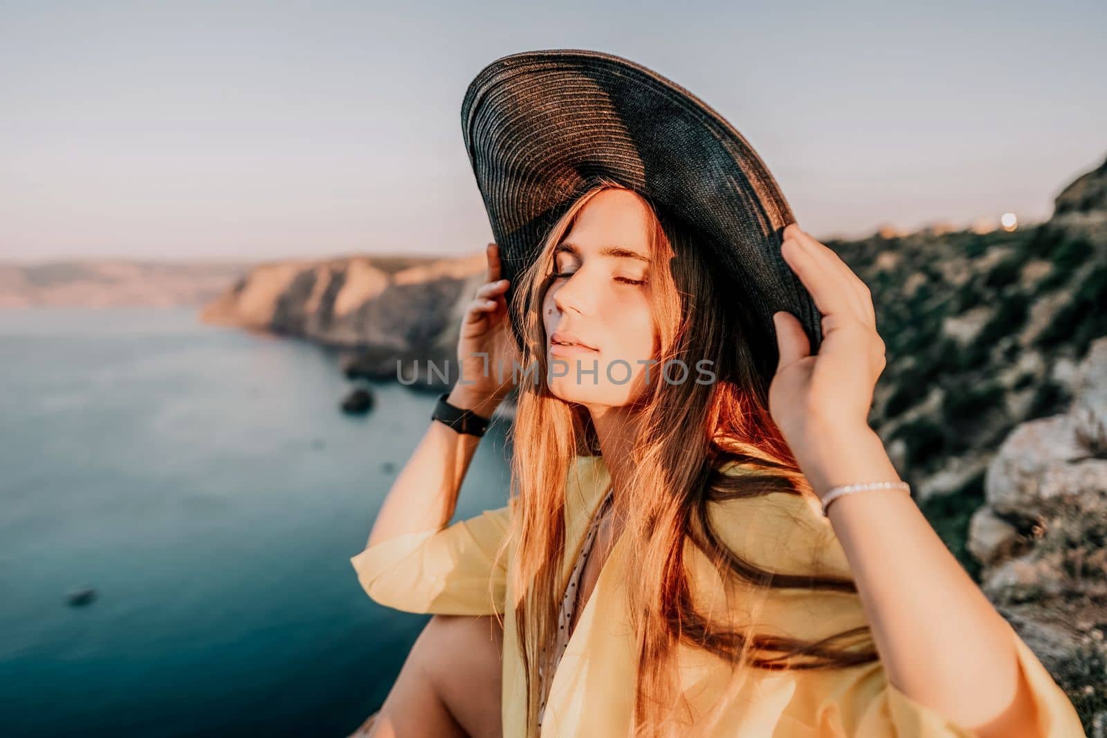 Portrait of happy young woman wearing summer black hat with large brim at beach on sunset. Closeup face of attractive girl with black straw hat. Happy young woman smiling and looking at camera at sea