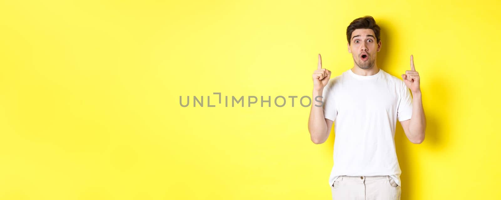 Surprised handsome guy in white t-shirt, pointing fingers up, interested about advertisement, standing against yellow background.