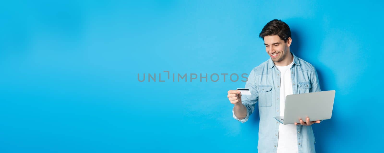 Young man order online, holding credit card and laptop, shopping in internet, standing against blue background.