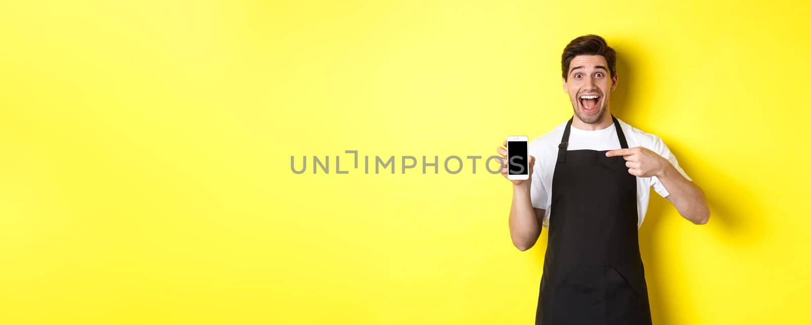 Handsome barista in black apron pointing finger at mobile screen, showing app and smiling, standing over yellow background.