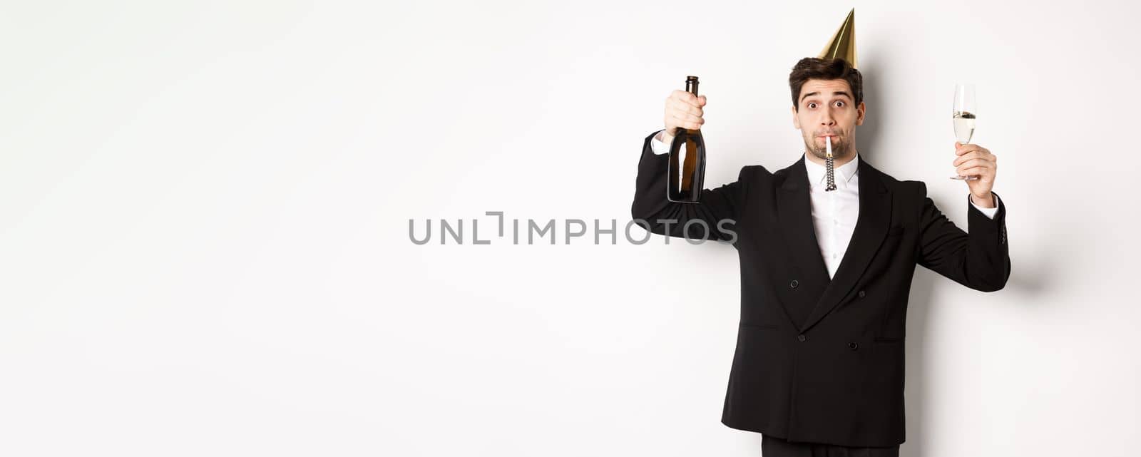 Concept of holidays and lifestyle. Handsome guy celebrating birthday, blowing party whistle and holding champagne, saying a toast, standing in suit over white background.