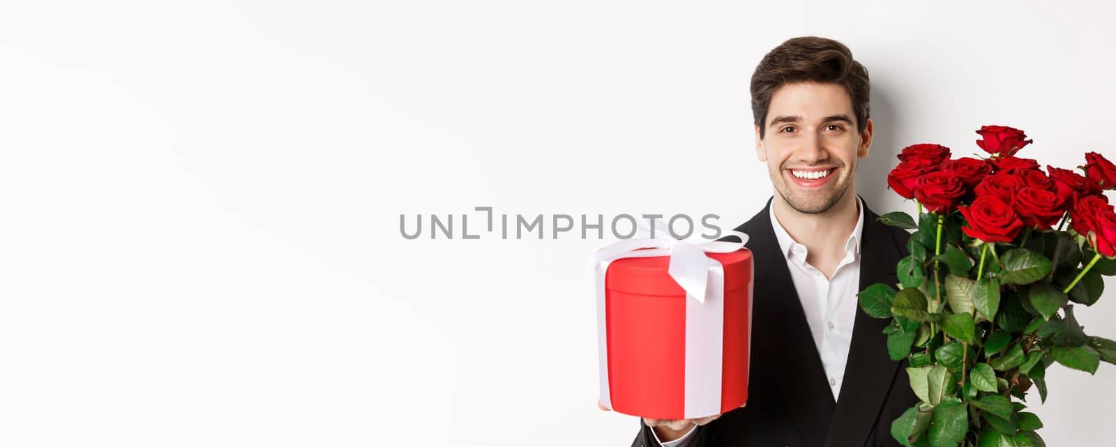 Close-up of handsome bearded man in suit, holding present and bouquet of red roses, smiling at camera, standing against white background by Benzoix