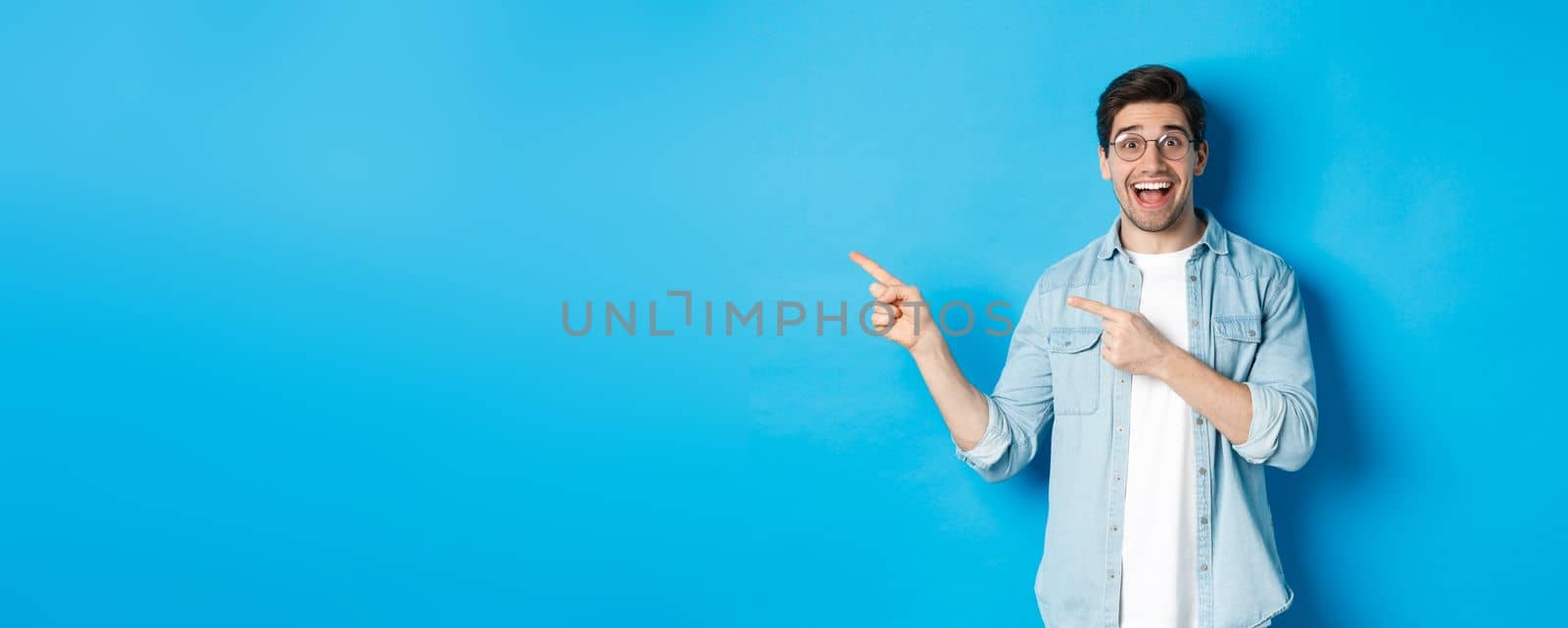 Excited handsome man in glasses pointing fingers left, looking happy while showing advertisement, demonstrating copy space on blue background by Benzoix