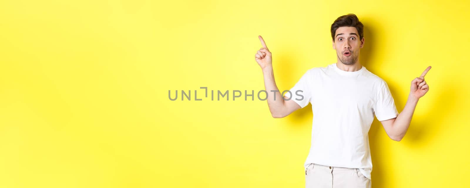 Handsome man pointing fingers sideways, showing two promos, standing over yellow background by Benzoix