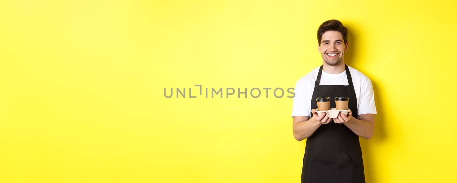 Handsome male barista serving takeaway coffee and smiling, bringing order, standing in black apron against yellow background by Benzoix