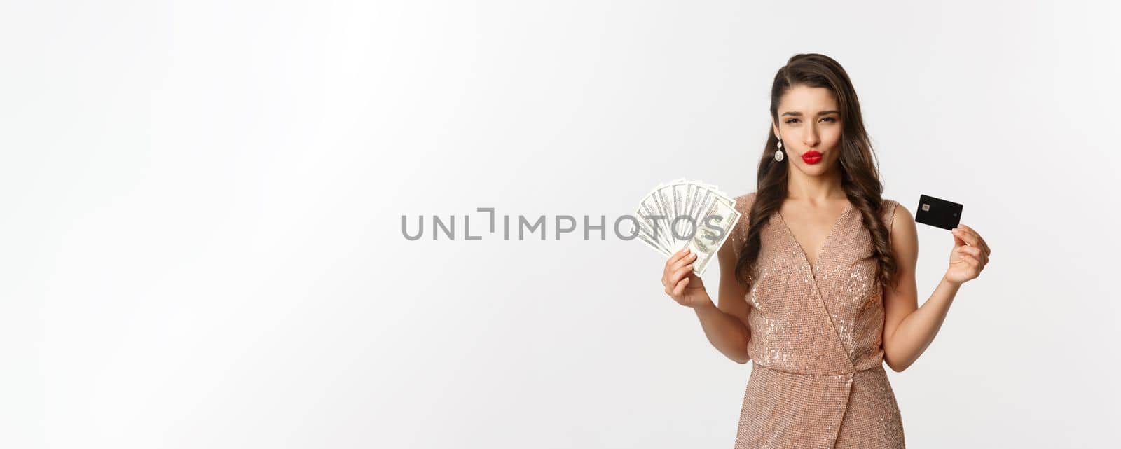 Shopping concept. Sassy woman in elegant dress showing credit card with money, looking pleased at camera, standing over white background by Benzoix