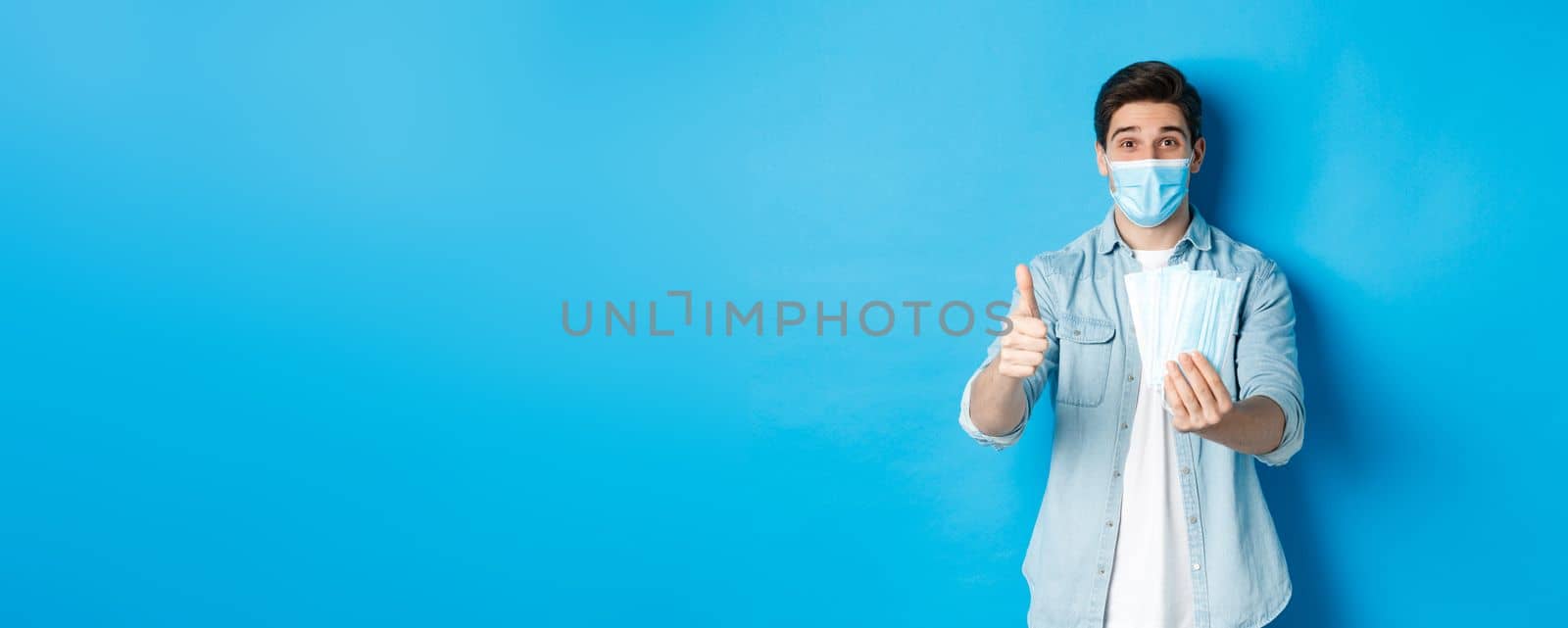 Concept of coronavirus, quarantine and social distancing. Young man showing medical masks and thumb up, standing against blue background by Benzoix