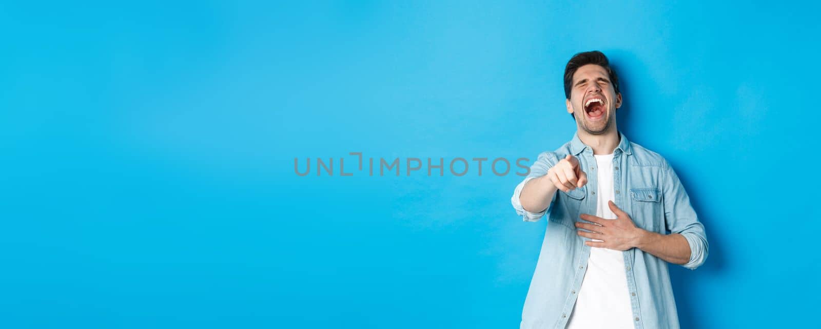 Adult man in casual outfit laughing out loud and pointing at you, looking at something funny, chuckling while standing against blue background by Benzoix