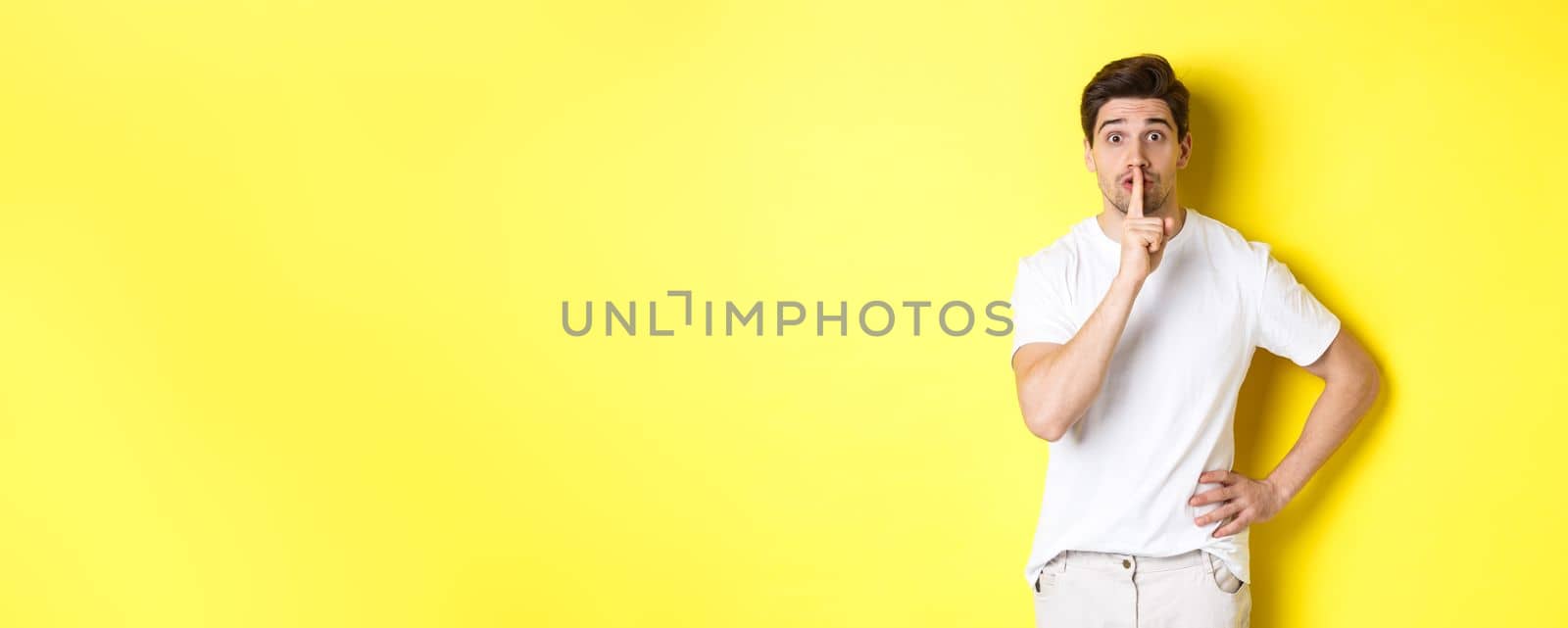 Image of excited guy planning surprise, shushing at camera, press finger to lip in hush sign, standing over yellow background.