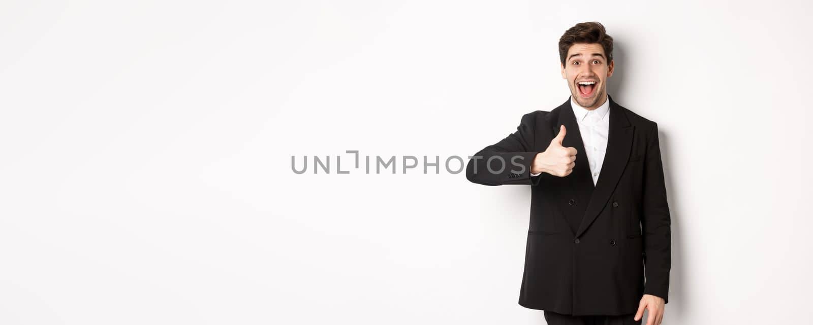 Portrait of amazed and impressed handsome man in black suit, showing thumb-up, praise awesome product, liking something good, standing against white background by Benzoix