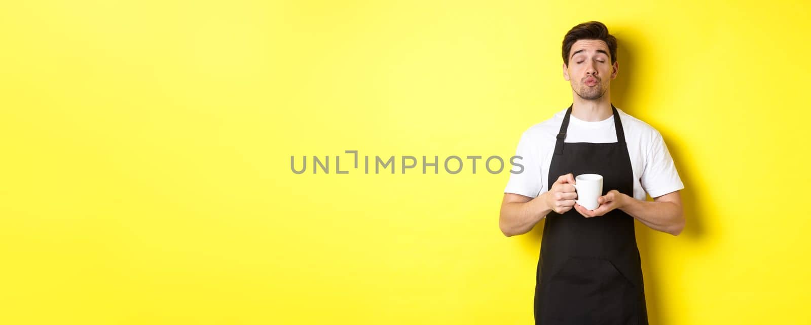 Man in black apron bringing cup of coffee and waiting for kiss, standing over yellow background.