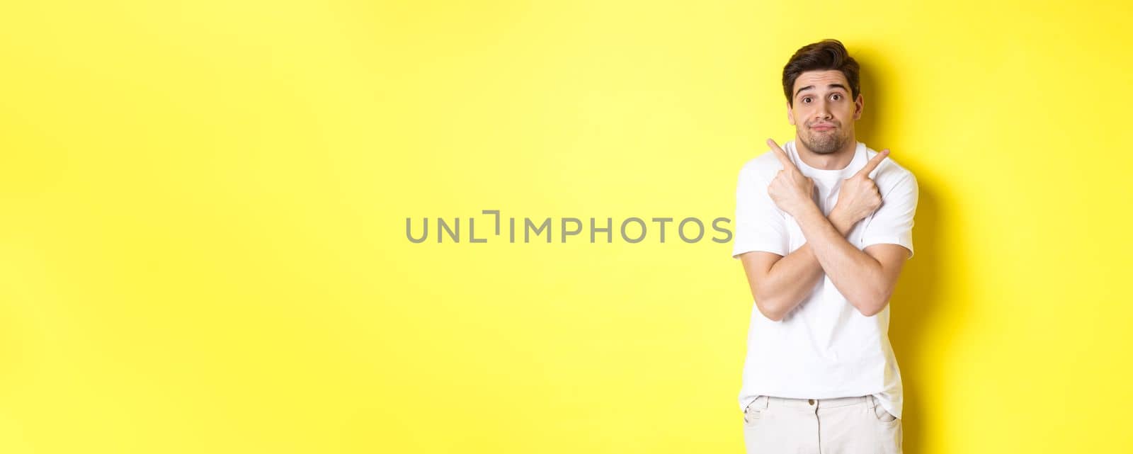 Indecisive man pointing fingers sideways, struggling to make decision, asking advice, standing over yellow background by Benzoix