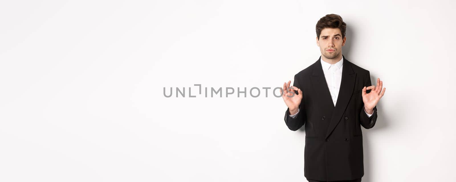 Concept of new year party, celebration and lifestyle. Portrait of confident good-looking man in black suit, showing okay sign and approve something, standing over white background by Benzoix