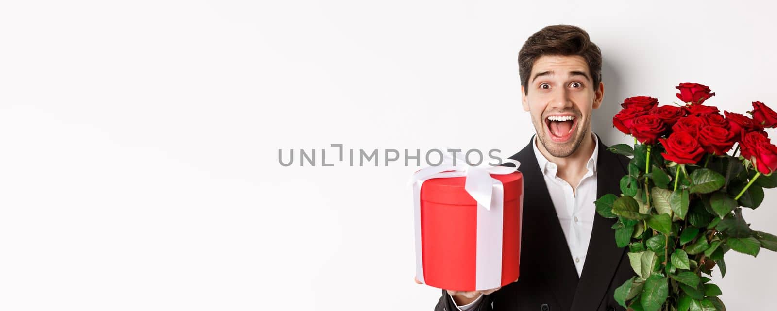 Close-up of handsome bearded man in suit, holding present and bouquet of red roses, smiling at camera, standing against white background by Benzoix