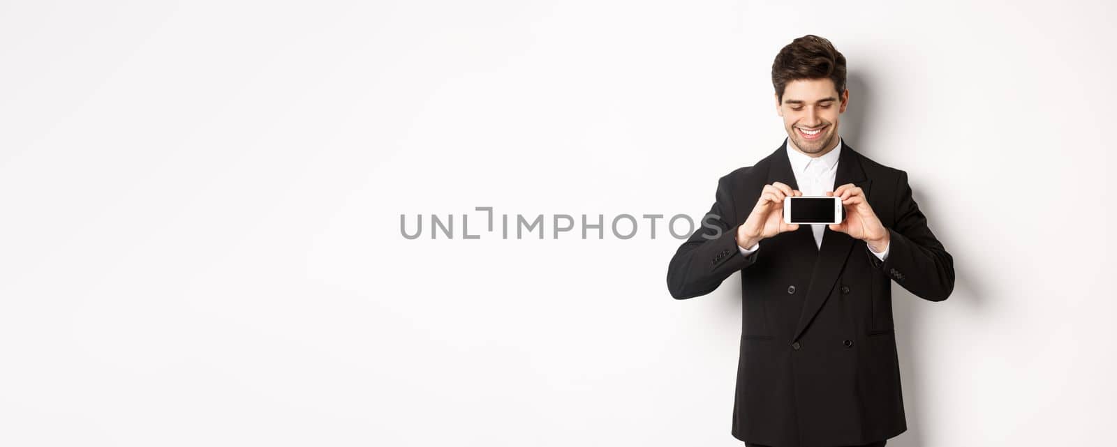 Portrait of handsome bearded man in black suit, showing you smartphone screen and smiling pleased, standing against white background.
