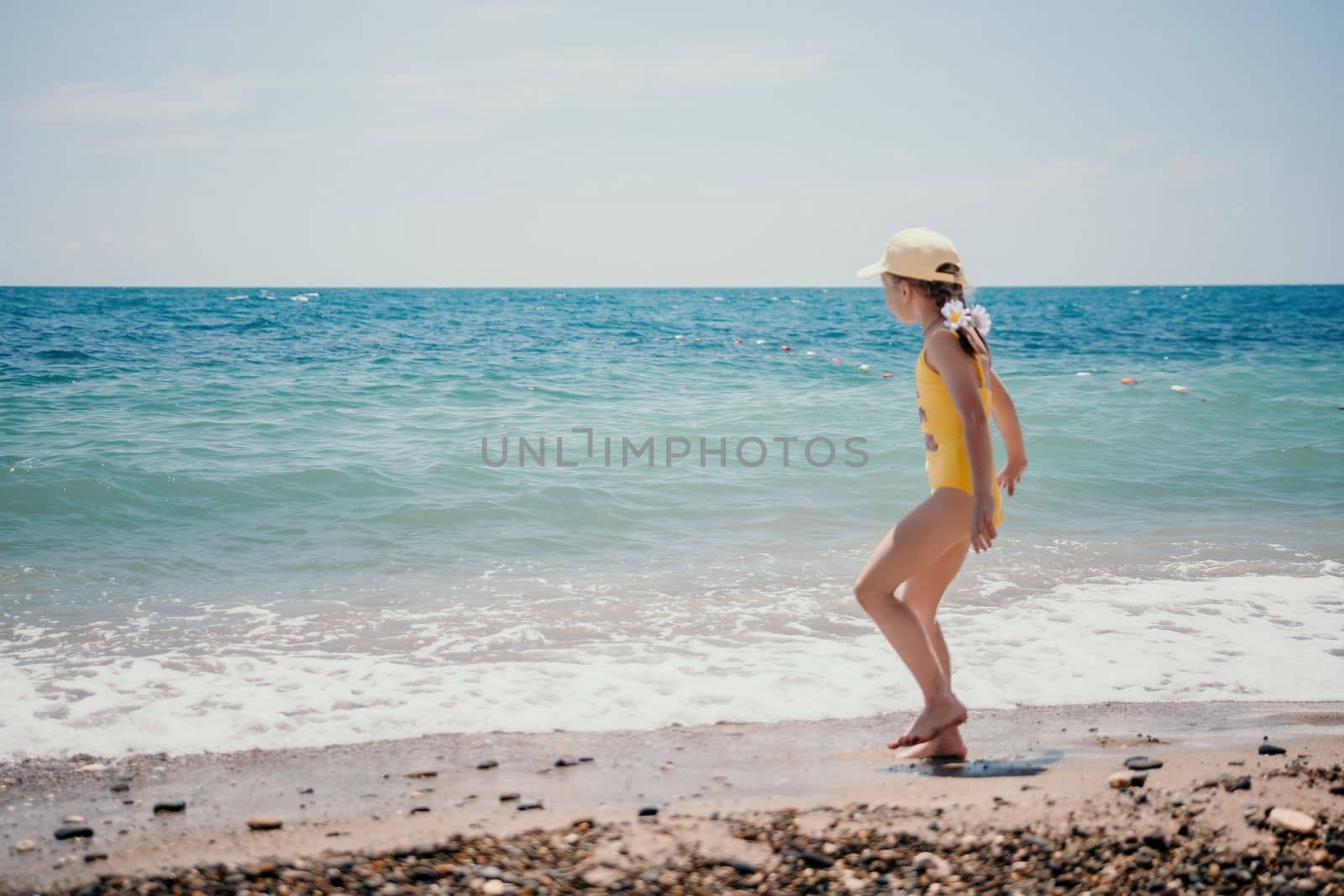 Cute little girl running along the seashore against a clear blue sea and rejoices in the rays of the summer sun. Beautiful girl in yellow swimsuit running and having fun on tropical beach