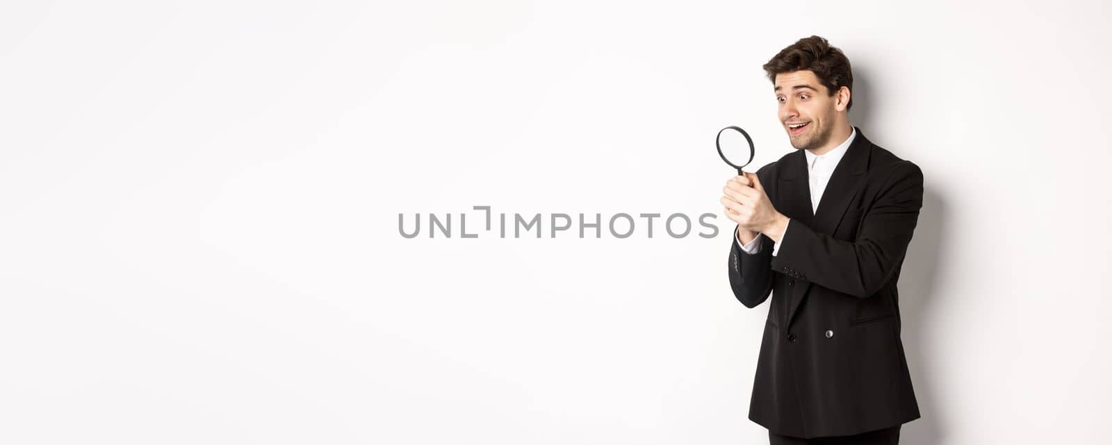 Image of handsome businessman in black suit, looking through magnifying glass, found something and smiling, standing over white background.