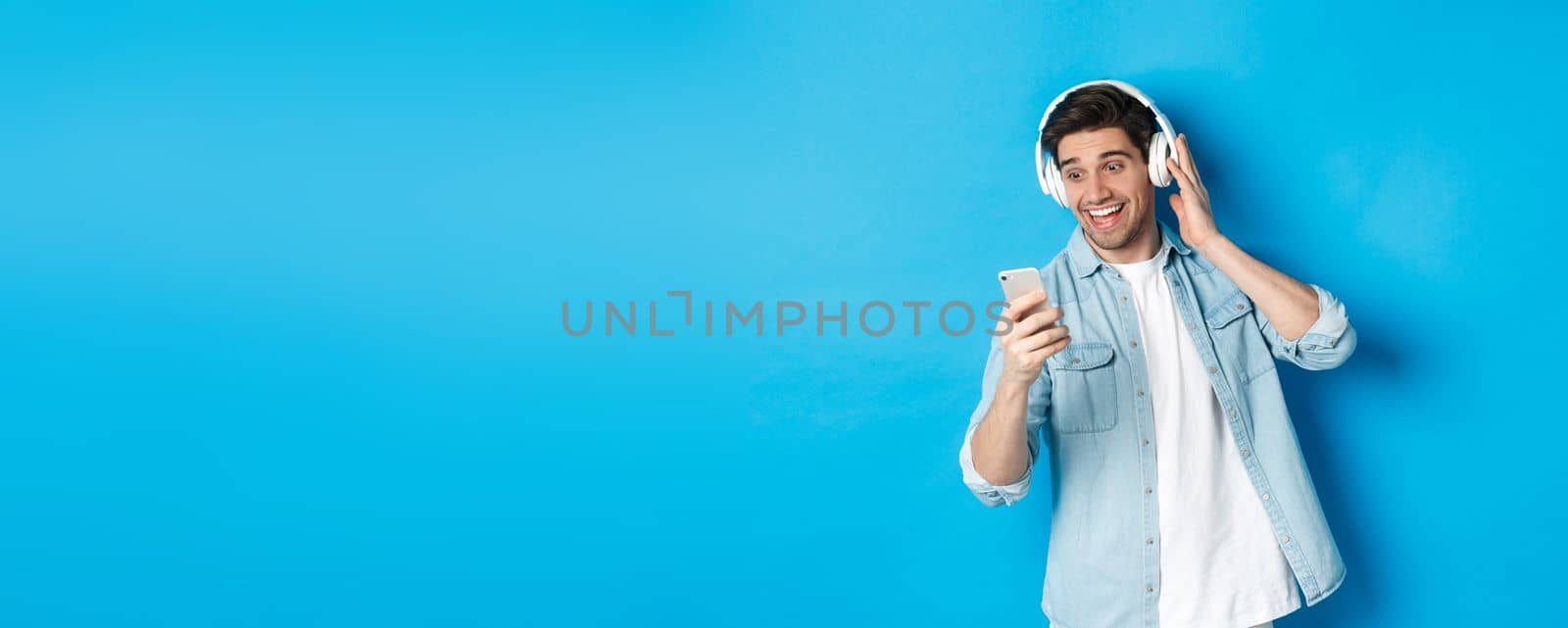 Happy man listening to music in headphones and reading message on smartphone, smiling excited, standing against blue background.