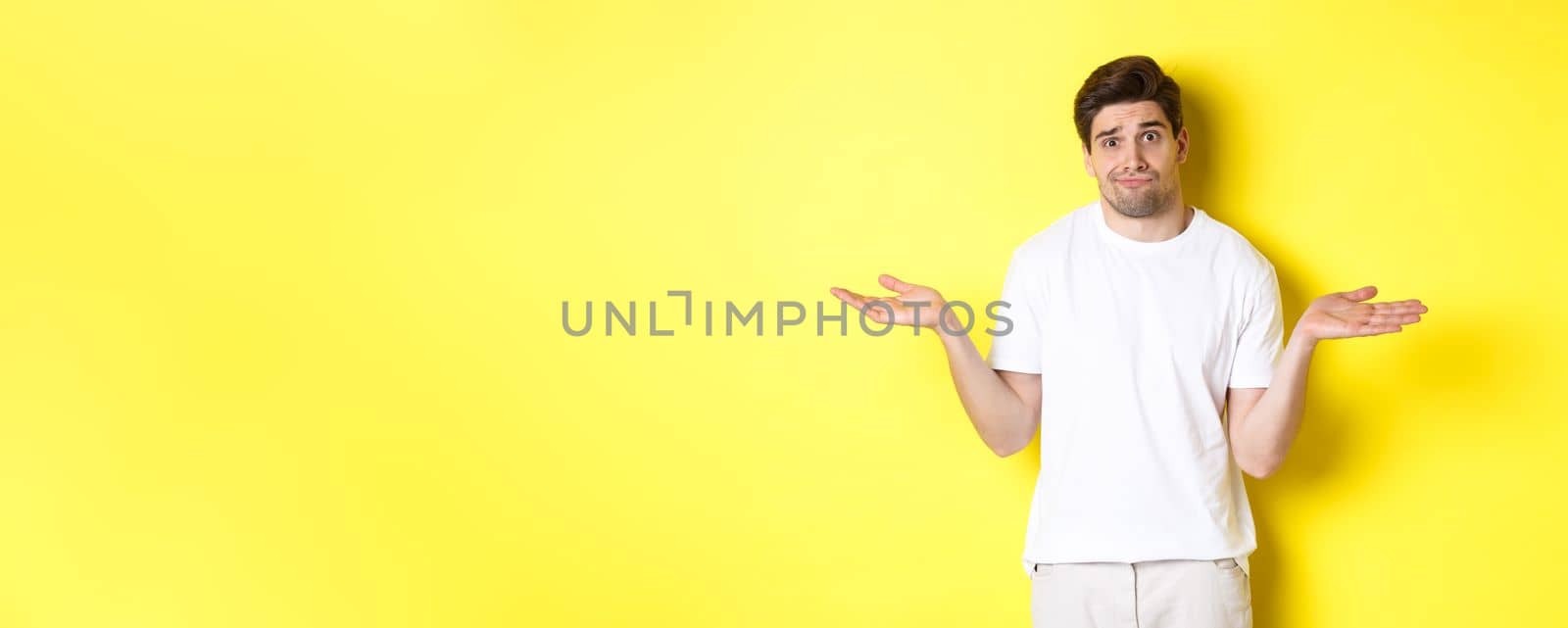 Clueless man in white t-shirt, shrugging and looking puzzled, dont know anything, standing over yellow background.