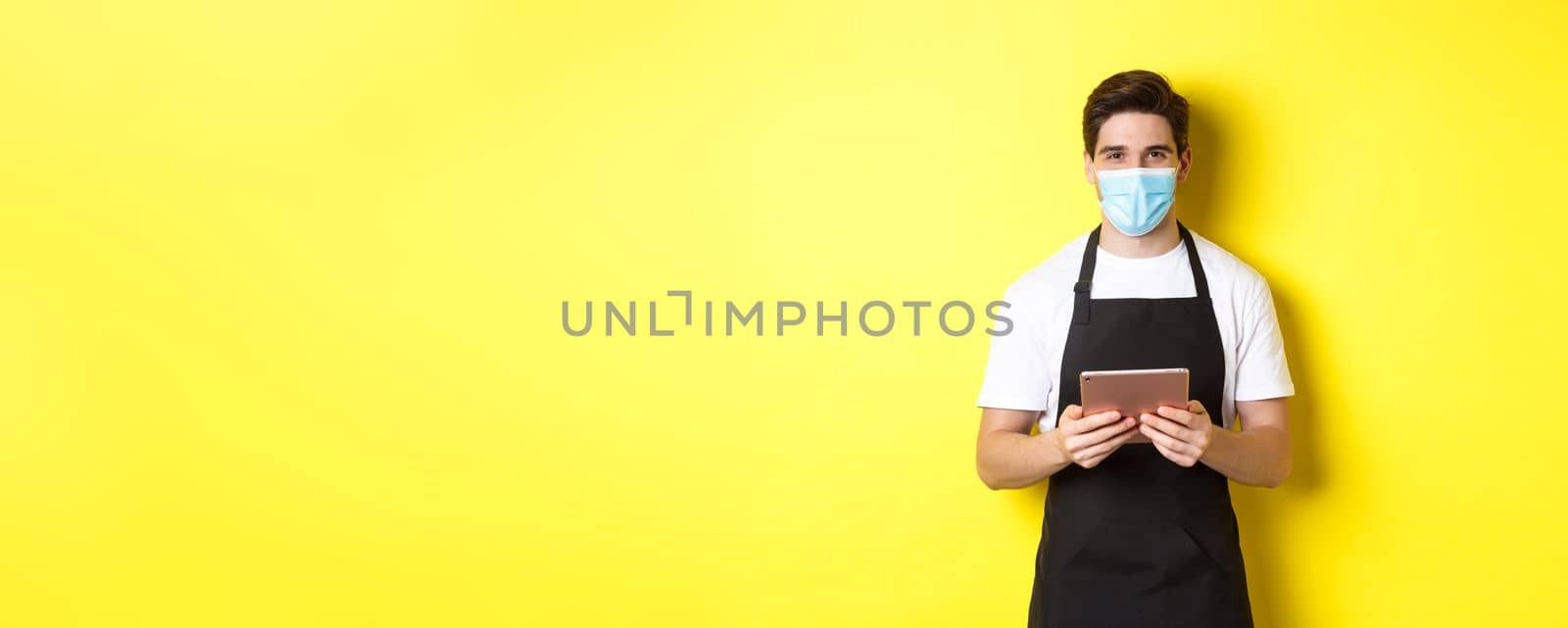 Concept of covid-19, small business and pandemic. Waiter in black apron and medical mask taking order, holding digital tablet, standing over yellow background.