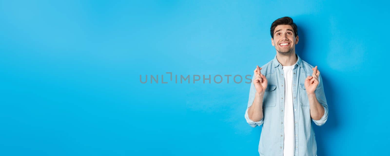 Lucky guy praying and making wish with crossed fingers, looking up with pleading face, standing against blue background.