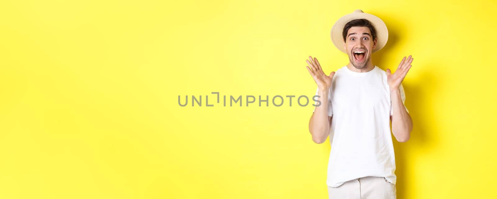 Concept of tourism and summer. Happy young man in straw hat looking amazed, reacting to surprise, standing over yellow background.