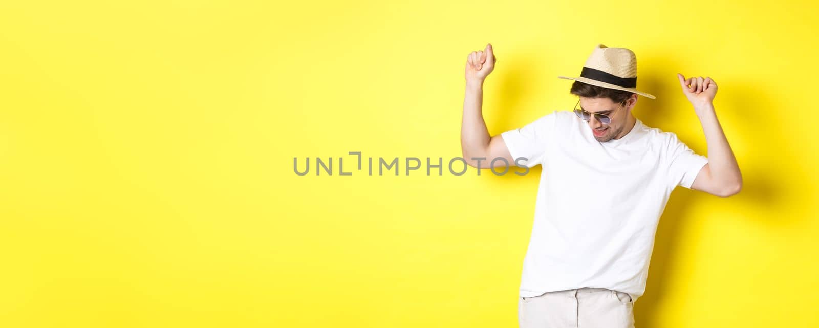 Tourism, travelling and holidays concept. Man tourist enjoying vacation, dancing in straw hat and sunglasses, posing against yellow background by Benzoix