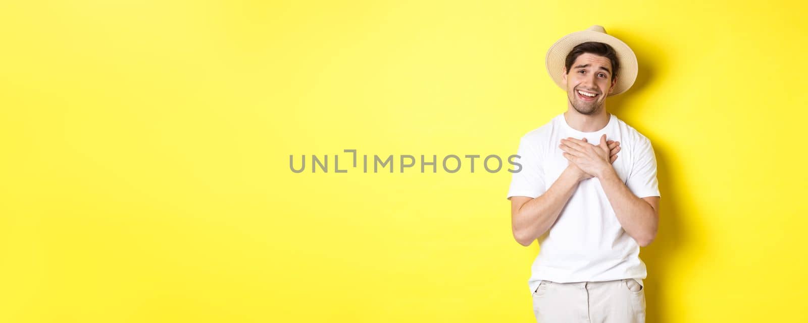 Concept of tourism and summer. Grateful guy in straw hat holding hands on heart, saying thank you and smiling with gratitude, standing against yellow background.