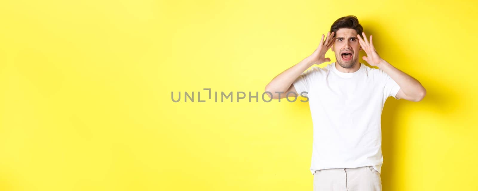 Frustrated guy looking alarmed, holding hands on head and feeling confused, panicking, standing over yellow background.