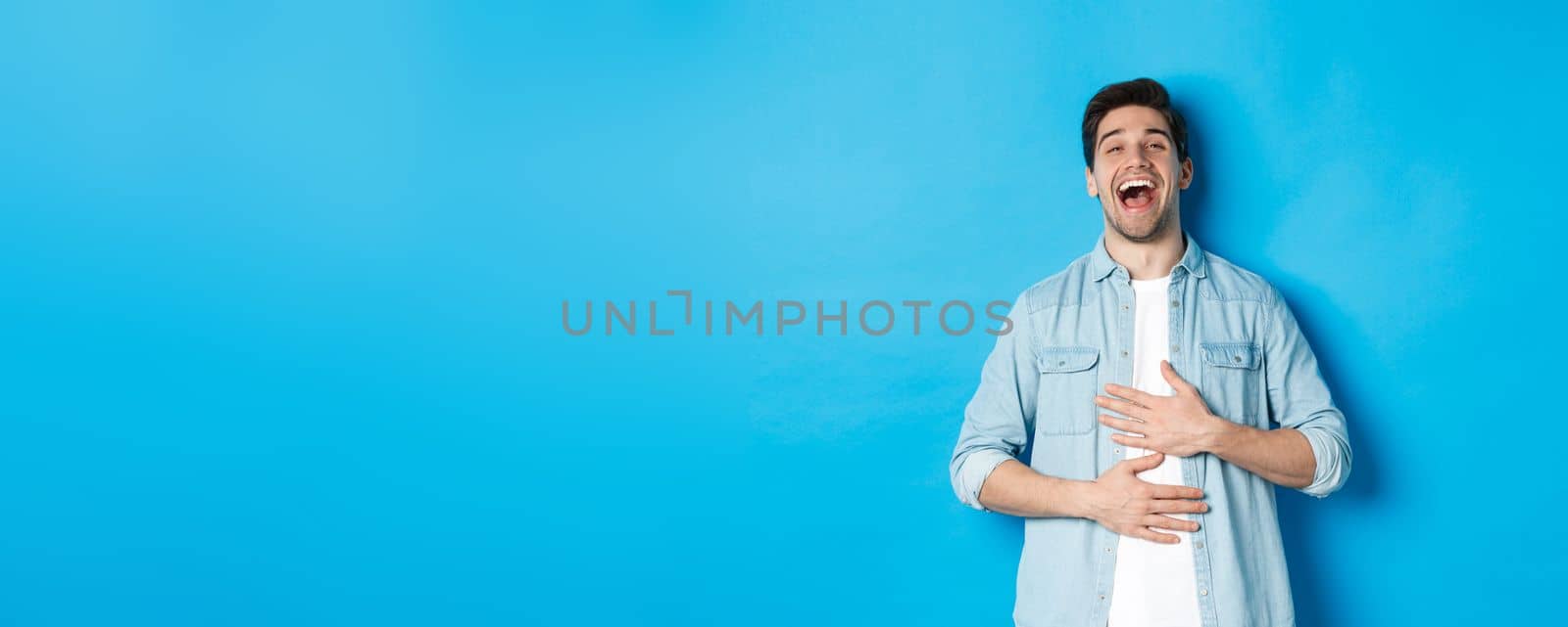Happy handsome 30 year old man touching belly, laughing out loud and looking at something funny, standing against blue background by Benzoix