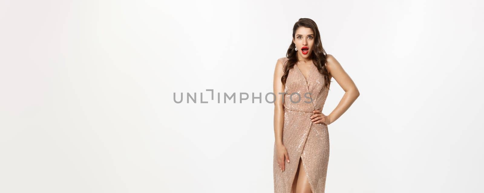 Christmas party and celebration concept. Full length of beautiful brunette woman looking surprised, wearing elegant dressed and gasping amazed at camera, white background.