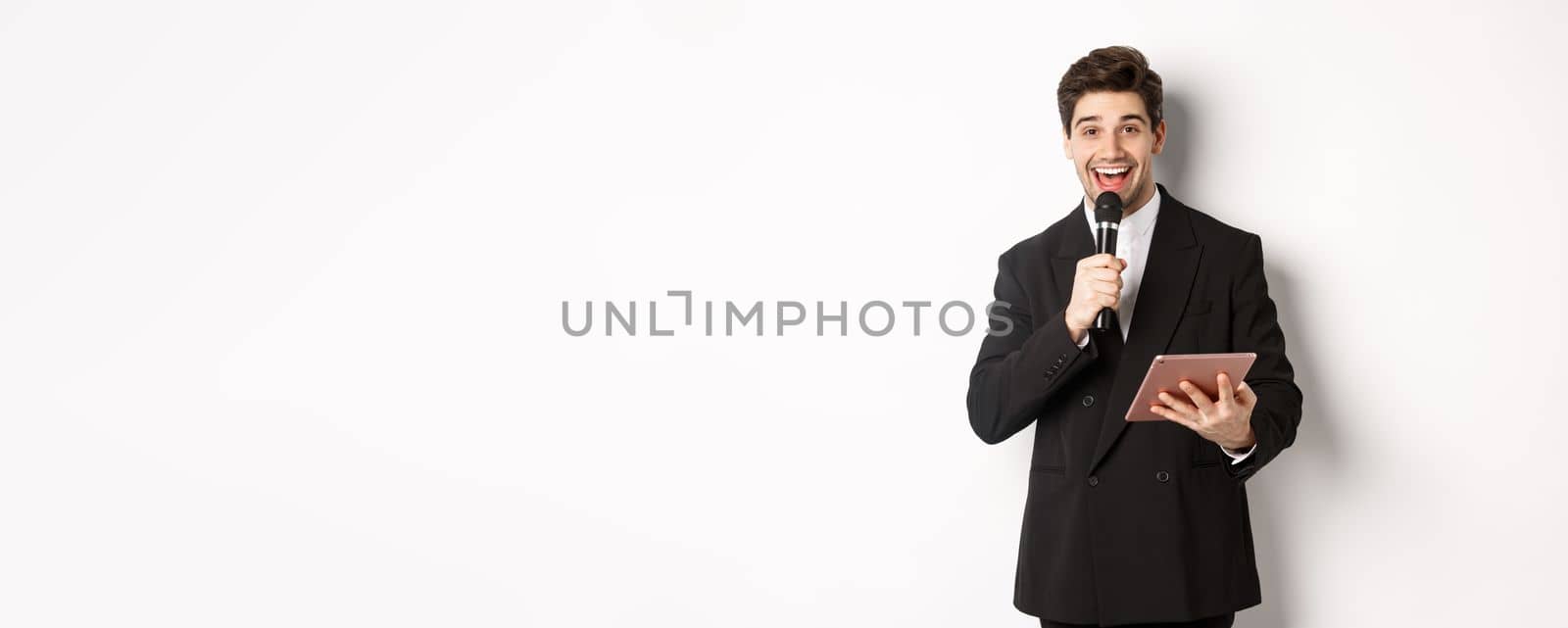 Image of handsome male performer, giving a speech, entertain people at party, holding microphone and digital tablet, standing over white background.