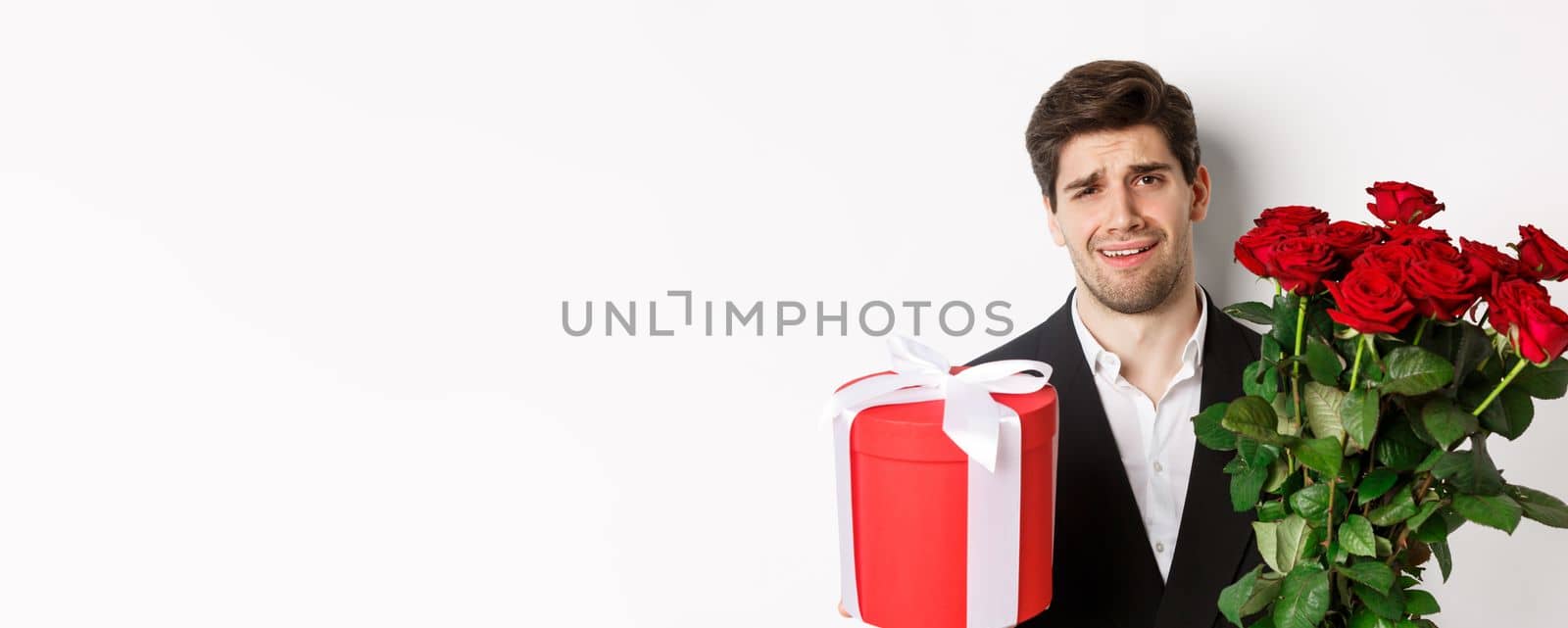 Close-up of skeptical man in suit, holding bouquet of red roses and a gift, standing reluctant against white background.