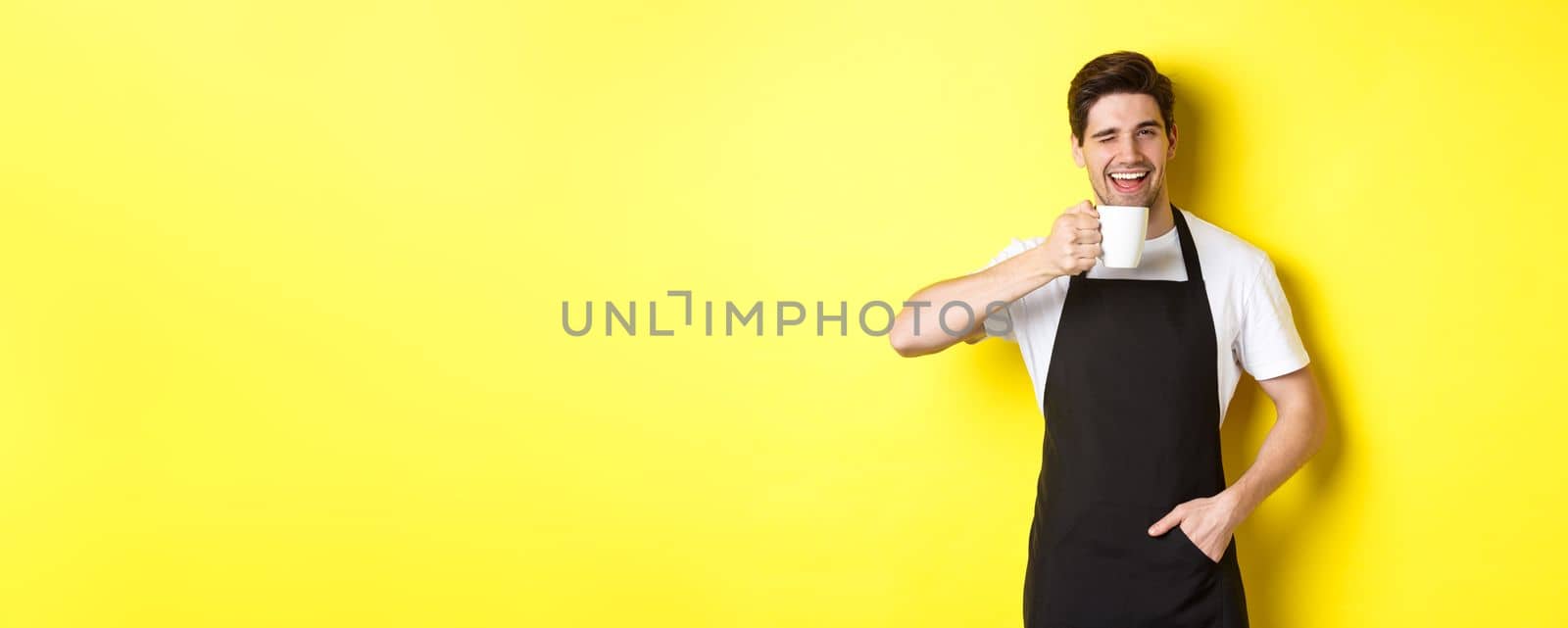 Handsome barista drinking cup of coffee and winking, inviting to cafe, standing over yellow background.