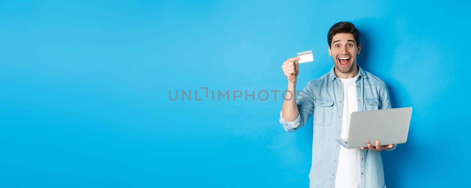 Excited man shop online, showing credit card and holding laptop, buying in internet, standing over blue background.