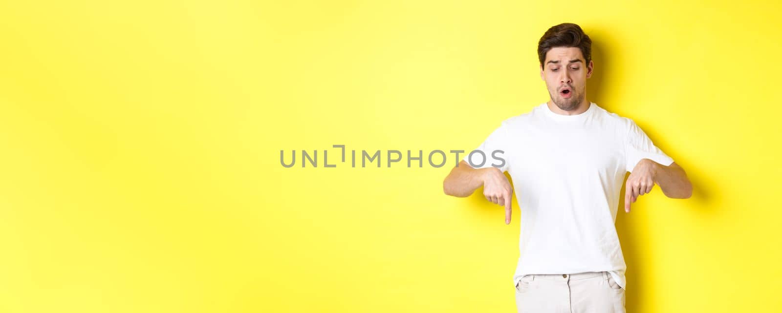 Worried guy pointing fingers and looking down, standing startled against yellow background.