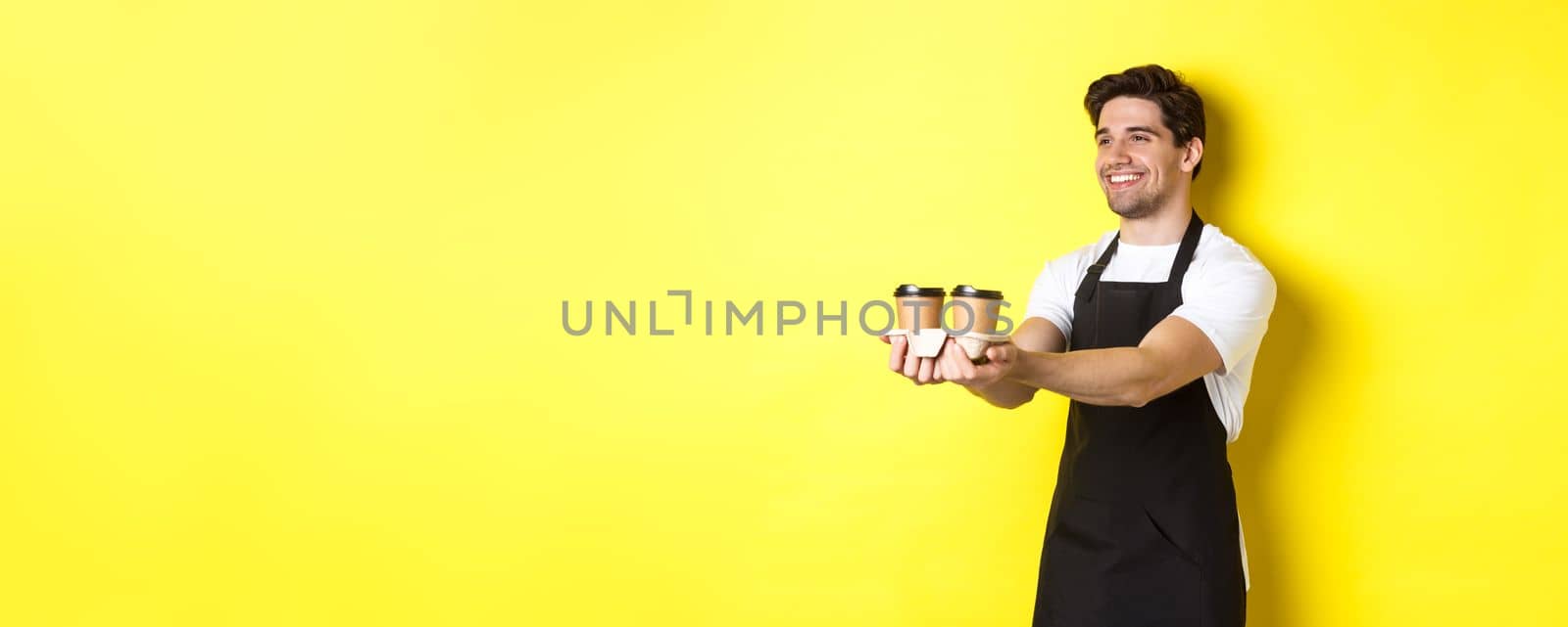 Friendly barista in black apron giving takeaway order, holding two cups of coffee and smiling, standing over yellow background by Benzoix