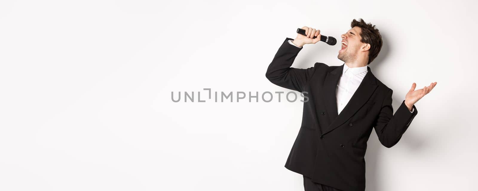 Portrait of handsome man singing a song with passion, standing in black suit, holding microphone and performning, posing over white background.