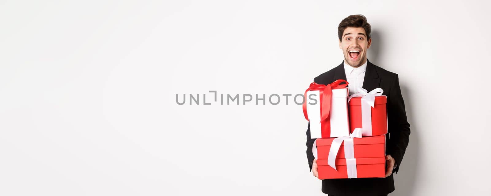 Concept of christmas holidays, celebration and lifestyle. Image of handsome amazed guy in suit, holding new year presents and smiling, standing against white background.