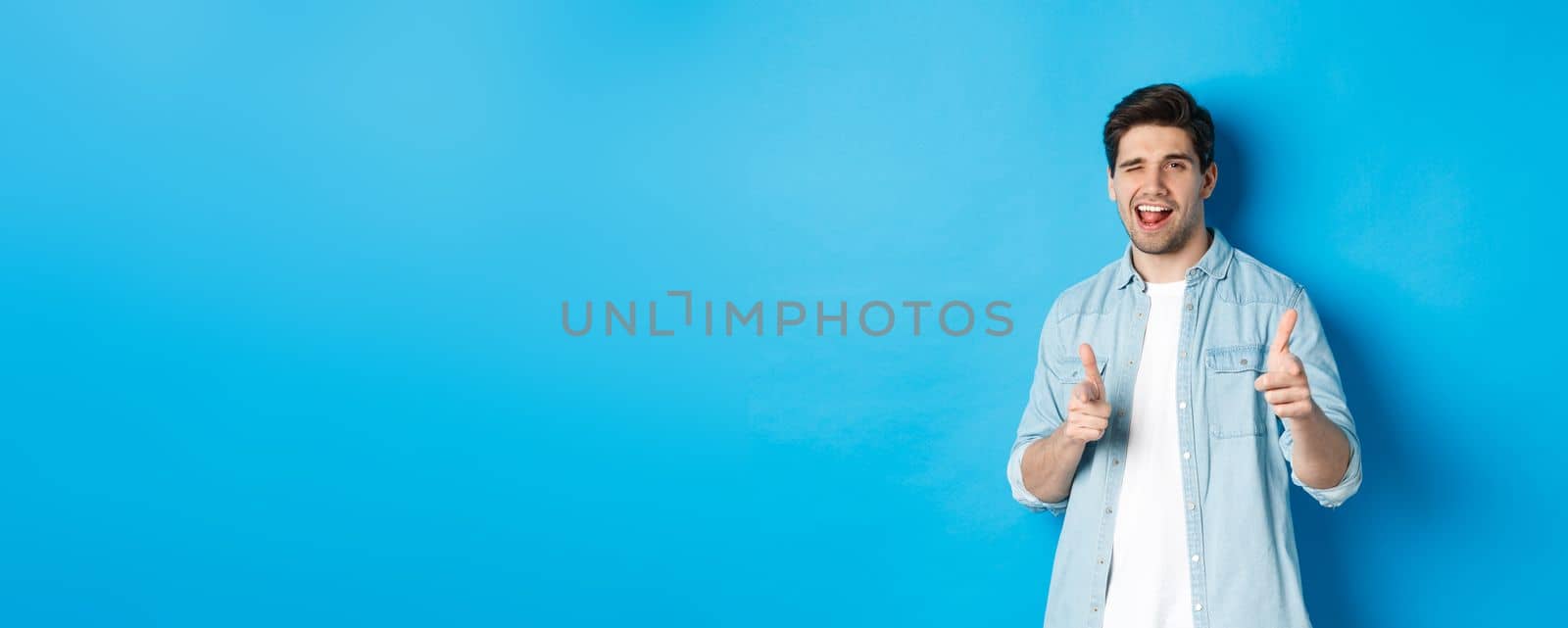 Confident adult man winking, pointing finger at you and winking, congratulating or praising person, standing against blue background by Benzoix