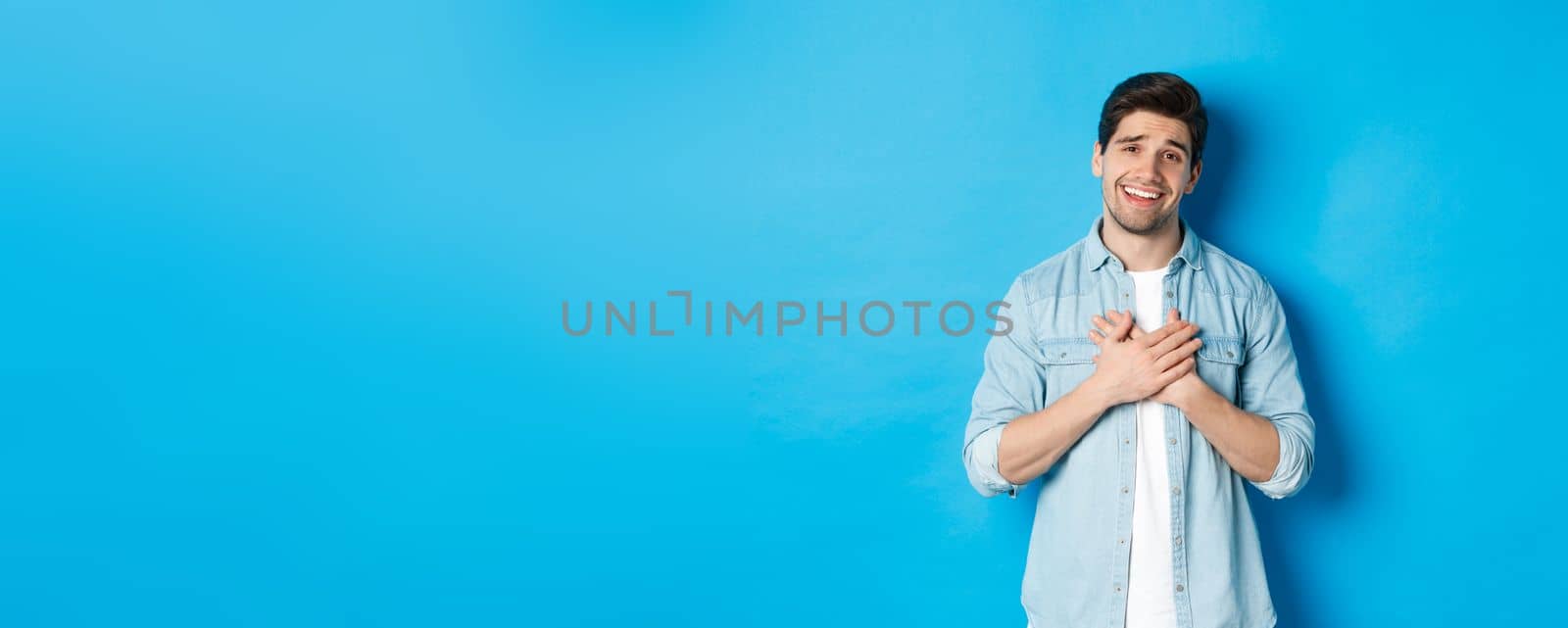 Portrait of flattered handsome man thanking you, holding hands on heart and smiling, looking grateful, standing over blue background.