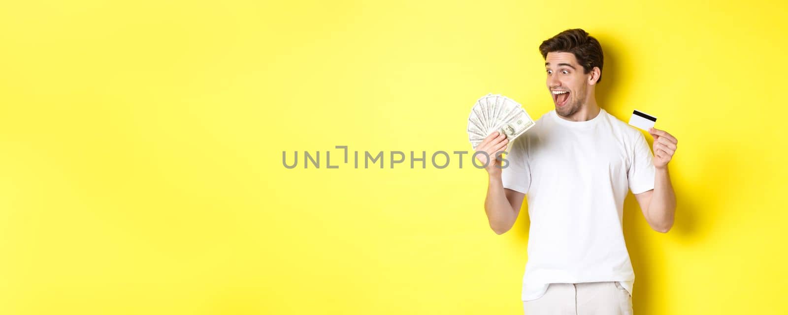 Cheerful guy looking at money, holding credit card, concept of bank credit and loans, standing over yellow background by Benzoix