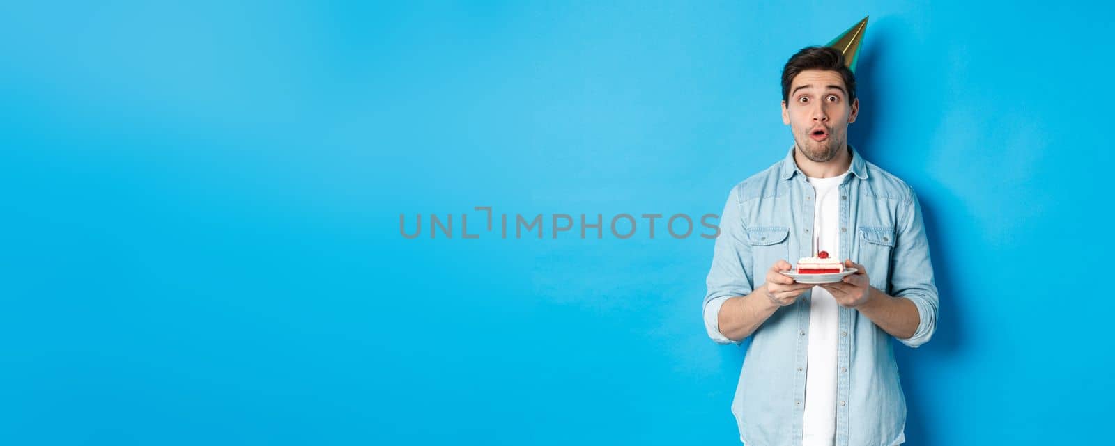 Handsome man in party cone holding birthday cake, looking surprised, standing over blue background by Benzoix