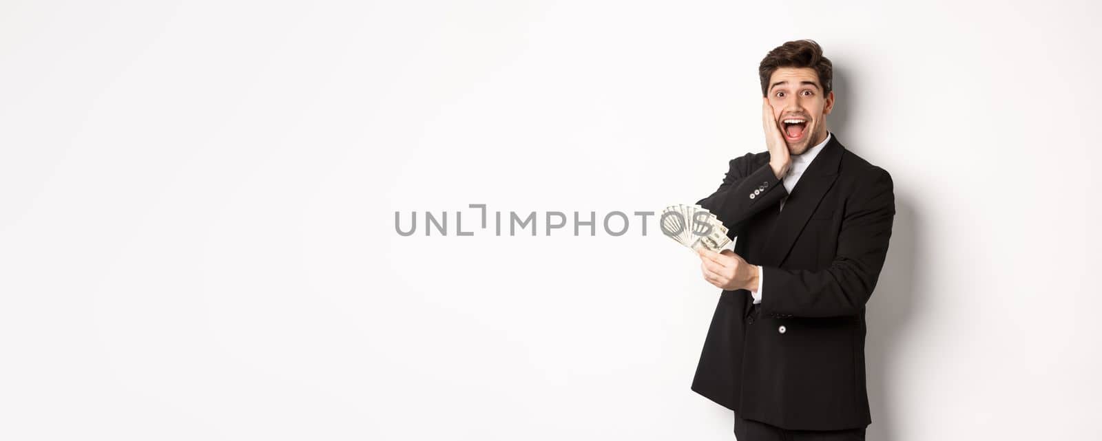 Image of rich and happy man in black suit, winning prize, holding money and looking excited at camera, standing over white background by Benzoix