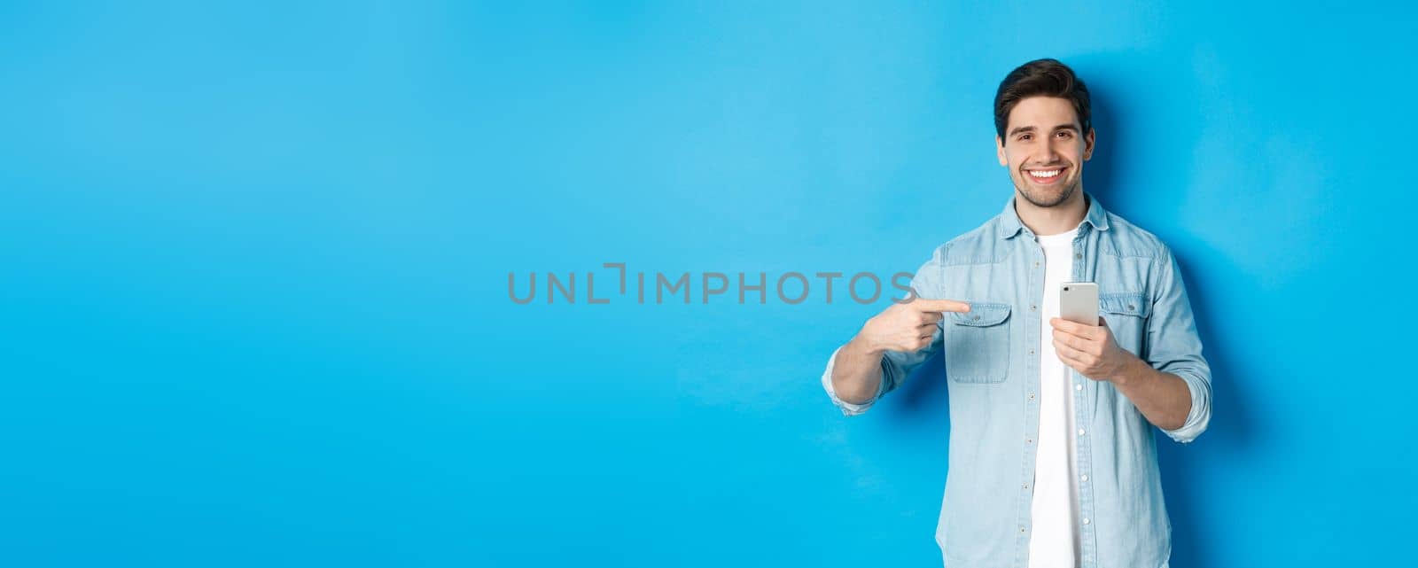 Concept of online shopping, applications and technology. Handsome man recommending app on smartphone, pointing at phone and smiling satisfied, standing over blue background by Benzoix