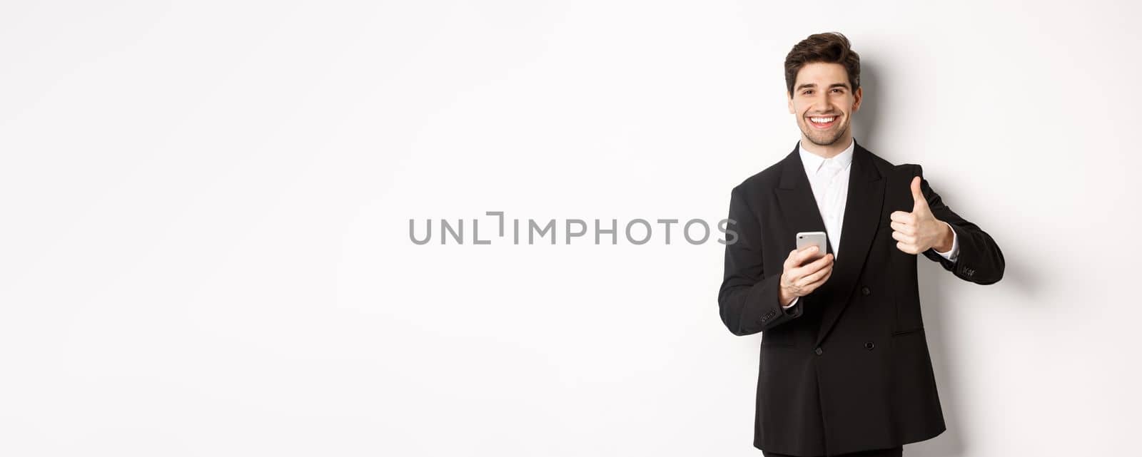 Handsome guy in trendy suit making compliment, showing thumbs-up while using mobile phone, recommending an app or online shop, standing over white background.