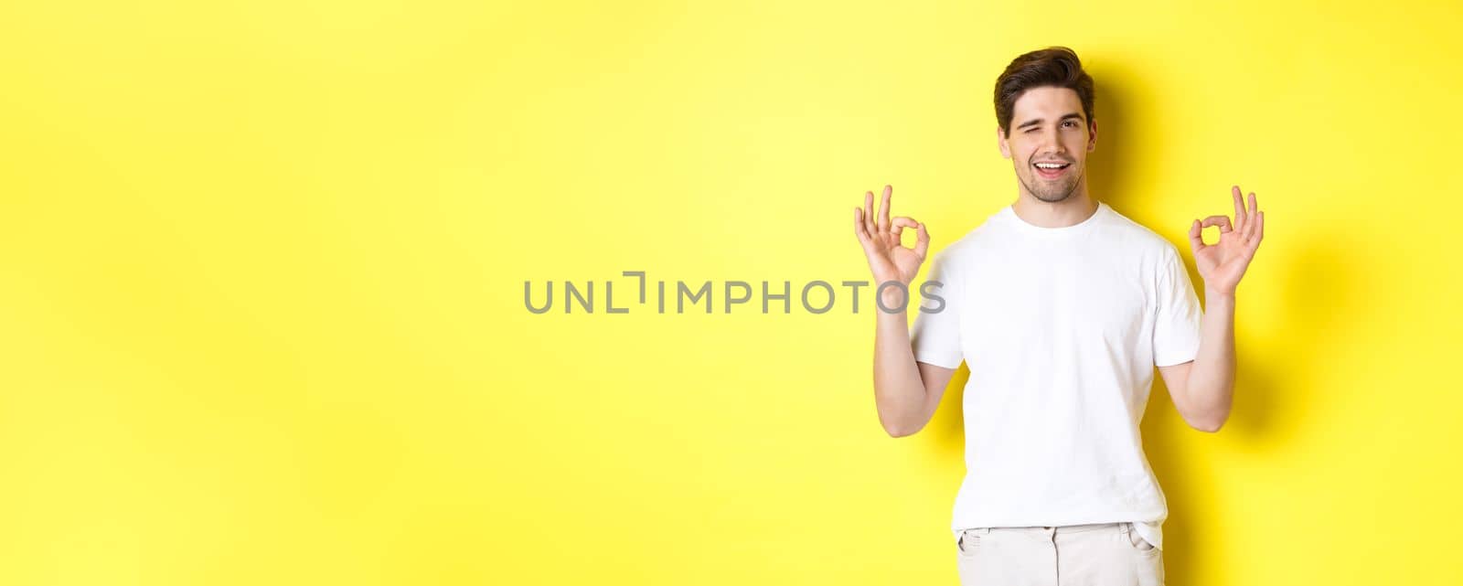 Confident handsome man winking, showing okay signs in approval, like something good, standing over yellow background.